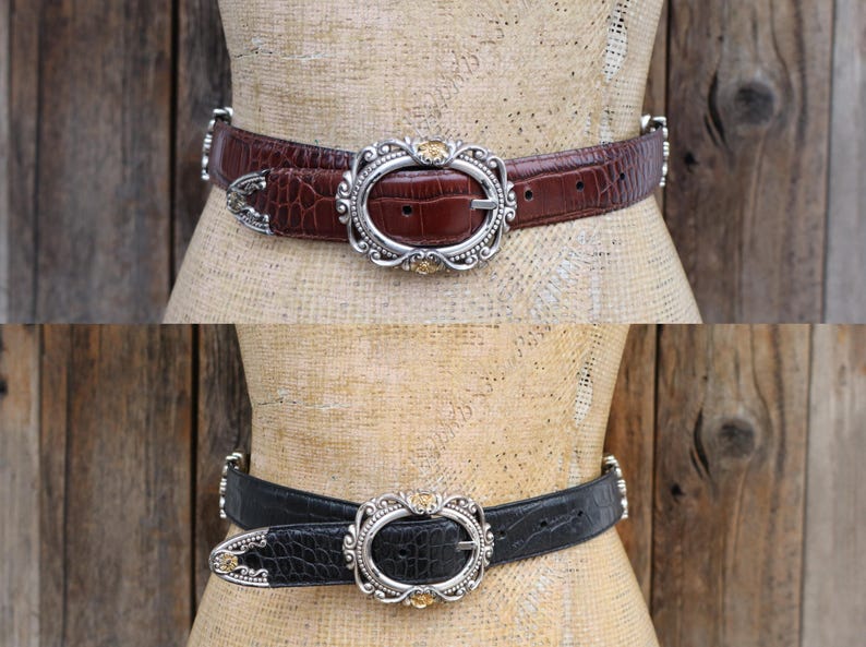 May include: Two belts are displayed, one brown and one black, each with a textured, crocodile-like pattern. Both belts feature an oval silver buckle with a decorative design and small gold-colored accents. The belts are displayed on a mannequin.