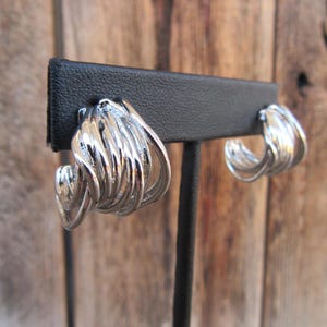 May include: A pair of silver hoop earrings with a twisted, layered design. The earrings are displayed on a black earring stand.