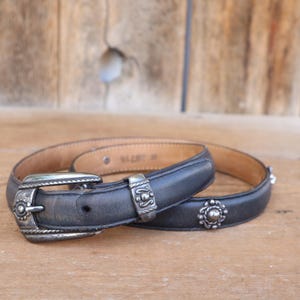 May include: A black leather belt with silver-toned metal buckle and decorative accents. The belt has a classic design with a medium width. The belt is lying on a wooden surface.