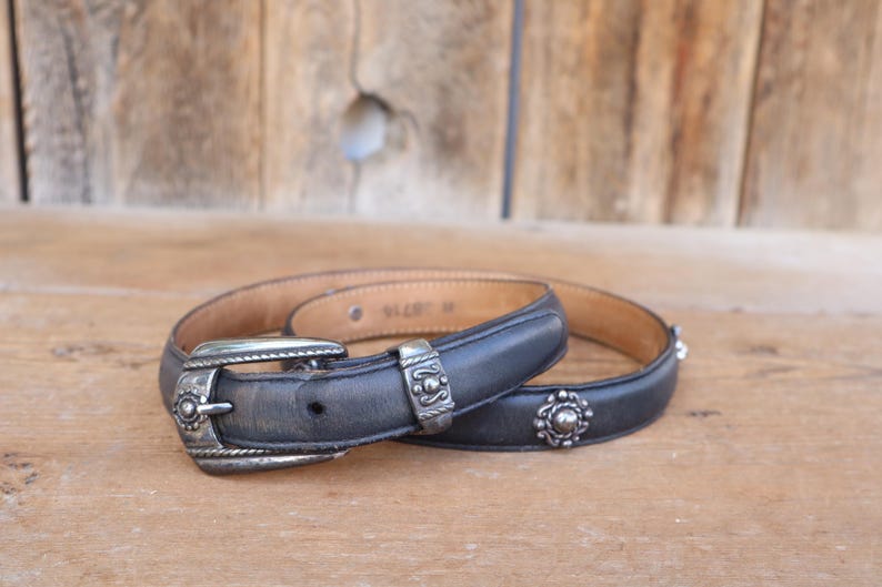May include: A black leather belt with silver-toned metal buckle and decorative accents. The belt has a classic design with a medium width. The belt is lying on a wooden surface.