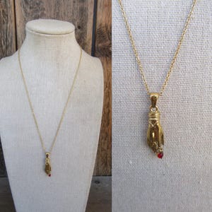 Puede incluir: Un collar de cadena dorada con un colgante con forma de mano que sostiene una piedra preciosa roja.