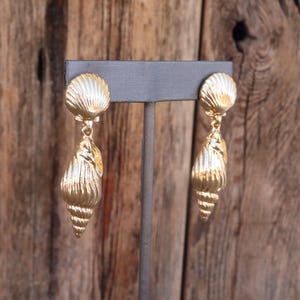 May include: A pair of gold-colored seashell earrings. Each earring features a round, ridged top and a dangling, textured conch shell design. The earrings are displayed on a gray stand against a wooden background.