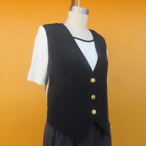 Peut inclure: Un gilet sans manches noir avec des boutons dor&eacute;s. Le gilet a une ourlet pointu et est port&eacute; sur un t-shirt blanc.