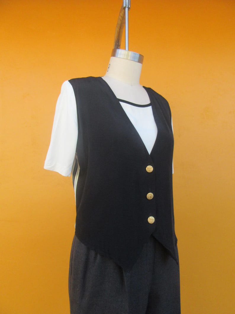 Peut inclure: Un gilet sans manches noir avec des boutons dor&eacute;s. Le gilet a une ourlet pointu et est port&eacute; sur un t-shirt blanc.
