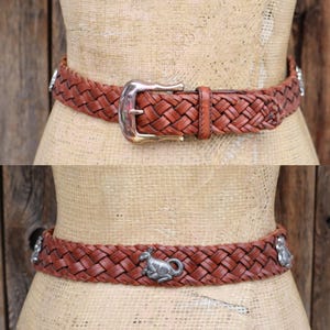 May include: A brown leather belt with a woven design. The belt has a silver buckle and decorative silver accents. The belt is displayed on a mannequin. The belt is approximately 3.8 cm wide.