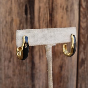 May include: A pair of gold hoop earrings with black enamel. The earrings are displayed on a white stand.