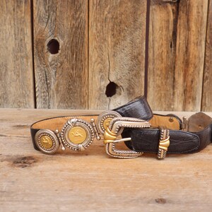 May include: A vintage black leather belt with ornate silver and gold-tone buckles and medallions. The belt features a textured leather design and decorative metal accents, showcasing a classic Western style.