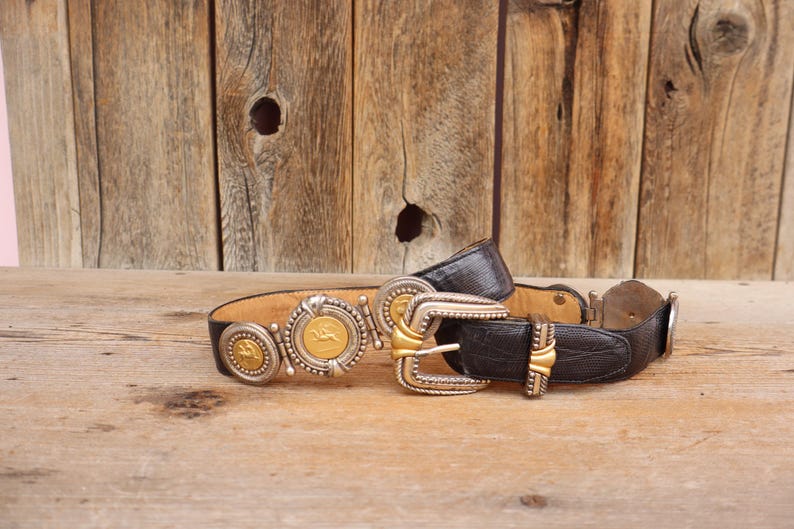 May include: A vintage black leather belt with ornate silver and gold-tone buckles and medallions. The belt features a textured leather design and decorative metal accents, showcasing a classic Western style.