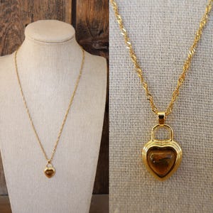 May include: A gold-toned necklace with a heart-shaped pendant. The pendant features a brown, heart-shaped stone set within a gold frame. The necklace is displayed on a mannequin and against a neutral background.