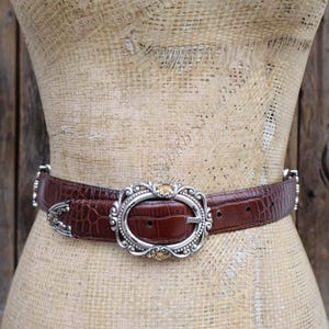 May include: A brown belt with a crocodile texture and a silver oval buckle with gold accents. The belt is displayed on a mannequin against a wooden background. The belt is approximately 1.5 inches wide.