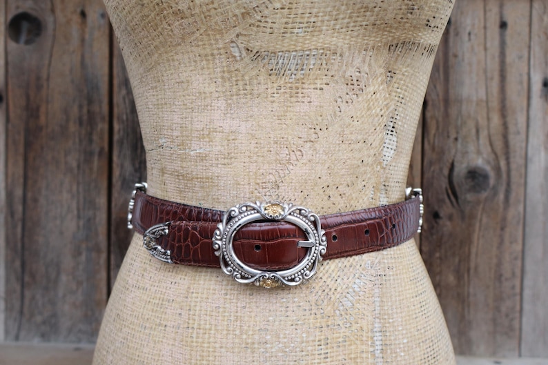 May include: A brown belt with a crocodile texture and a silver oval buckle with gold accents. The belt is displayed on a mannequin against a wooden background. The belt is approximately 1.5 inches wide.