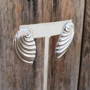 May include: A pair of silver-toned earrings. Each earring has a curved, elongated shape with a spiral design at the top and ridged detailing. The earrings are displayed on a white fabric stand against a wooden background.