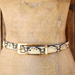 May include: A snakeskin-patterned belt with a gold-colored rectangular buckle. The belt is wrapped around a mannequin. The background is a wooden wall.
