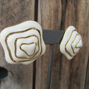 May include: A pair of white and gold earrings with a swirl design. The earrings are on a black stand.