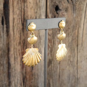 May include: Gold-toned seashell earrings. Each earring features three seashell charms of varying sizes, suspended from a small stud. The earrings are displayed on a gray stand against a wooden background. The seashells have a textured, ridged design.