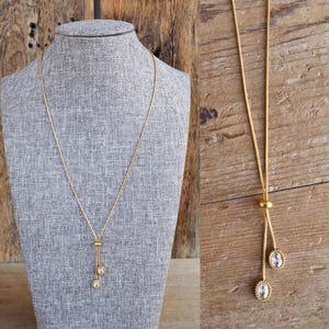 May include: A gold-toned necklace with a delicate chain and two oval pendants. The pendants have a clear, sparkling center surrounded by a gold frame. The necklace is displayed on a gray mannequin head against a wooden background.