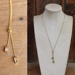 May include: A gold-toned necklace with a delicate chain and two rectangular, clear crystal pendants. The necklace is displayed on a mannequin and also shown on a wooden surface. The design features a sliding bead for adjustable length, offering a modern and elegant look.