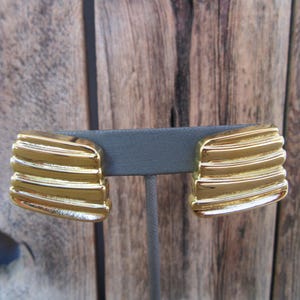 May include: A pair of gold-tone clip-on earrings with a ridged design. The earrings are displayed on a black stand.