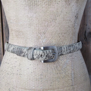 Op de afbeelding: Een lichtbruine leren riem met een zilveren gesp. De riem heeft een slangenhuidpatroon.