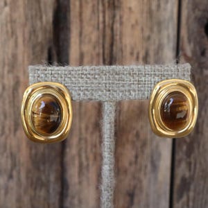 K&ouml;nnte beinhalten: Ein Paar goldfarbene, ovale Clip-Ohrringe. Jeder Ohrring hat einen braunen, ovalen Stein mit Tigeraugen-Effekt, der in einen goldenen Rahmen gefasst ist. Die Ohrringe werden auf einem neutralfarbenen Ohrringst&auml;nder vor einem Holzgrund pr&auml;sentiert.