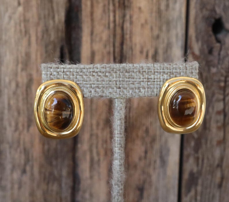 K&ouml;nnte beinhalten: Ein Paar goldfarbene, ovale Clip-Ohrringe. Jeder Ohrring hat einen braunen, ovalen Stein mit Tigeraugen-Effekt, der in einen goldenen Rahmen gefasst ist. Die Ohrringe werden auf einem neutralfarbenen Ohrringst&auml;nder vor einem Holzgrund pr&auml;sentiert.