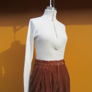 May include: A white ribbed turtleneck sweater with a zipper closure and long sleeves, paired with brown corduroy pants.