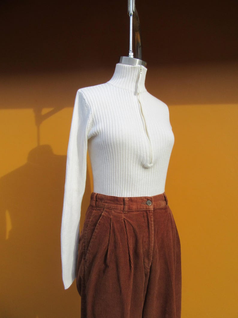 May include: A white ribbed turtleneck sweater with a zipper closure and long sleeves, paired with brown corduroy pants.