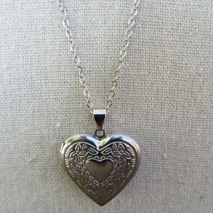 May include: Silver-colored heart-shaped locket necklace. The locket has an ornate floral design around a smaller heart in the center. The chain is a delicate silver color.