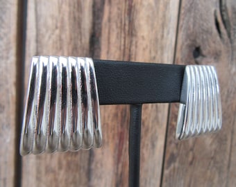 May include: A pair of silver earrings with a ridged, textured design. The earrings are displayed on a black stand.