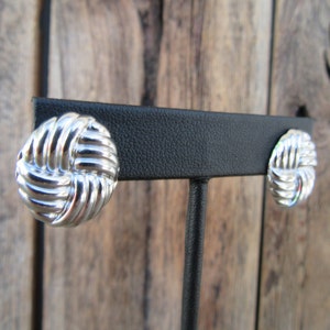 May include: A pair of silver knot-shaped earrings on a black display stand. The earrings are round and have a textured surface.