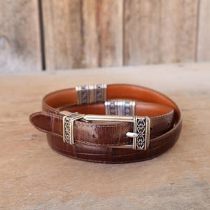 May include: A brown leather belt with a crocodile pattern. The belt has a silver-colored buckle and decorative accents. The belt is rolled up on a wooden surface.