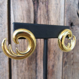 May include: A pair of gold-tone earrings with a wave-like design. The earrings are displayed on a black stand.