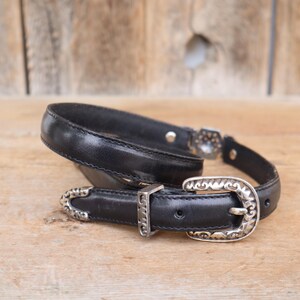 May include: Black leather belt with a silver-colored buckle and accents. The belt has a classic design with a decorative buckle and a matching keeper. The belt is displayed on a wooden surface.