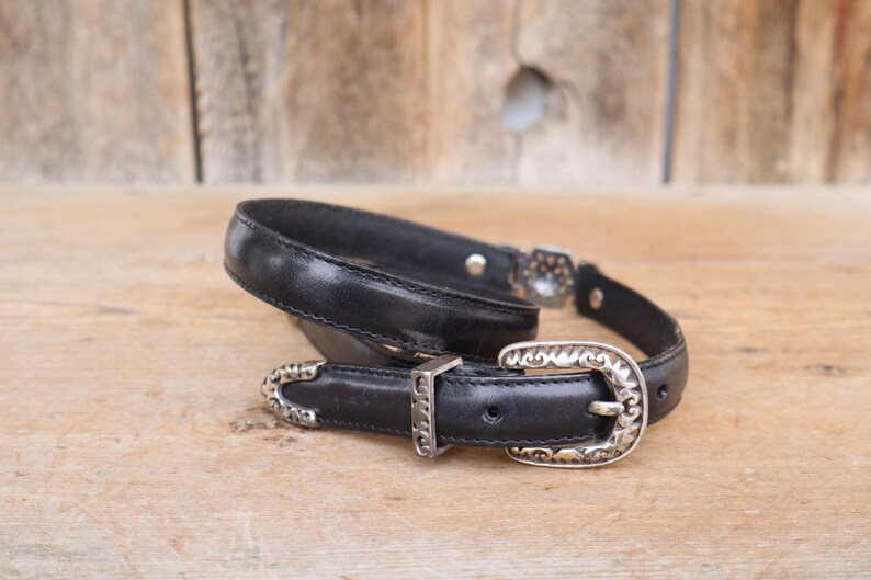 May include: Black leather belt with a silver-colored buckle and accents. The belt has a classic design with a decorative buckle and a matching keeper. The belt is displayed on a wooden surface.
