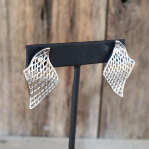 May include: A pair of silver-toned earrings with a unique openwork design. Each earring has a curved, diamond-like shape with a grid pattern, creating a textured, airy appearance. The earrings are displayed on a black stand against a blurred wooden background.