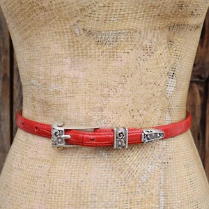 Puede incluir: Un cintur&oacute;n rojo con un patr&oacute;n texturizado y hebilla y detalles de metal plateado. El cintur&oacute;n se muestra en un maniqu&iacute;. La hebilla y los detalles tienen un dise&ntilde;o ornamentado.