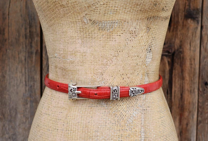 Puede incluir: Un cintur&oacute;n rojo con un patr&oacute;n texturizado y hebilla y detalles de metal plateado. El cintur&oacute;n se muestra en un maniqu&iacute;. La hebilla y los detalles tienen un dise&ntilde;o ornamentado.