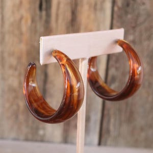 May include: A pair of C-shaped hoop earrings featuring a translucent, polished tortoiseshell pattern in shades of deep amber and dark brown. The earrings have a smooth, rounded profile and are displayed on a light-coloured T-shaped jewellery stand.
