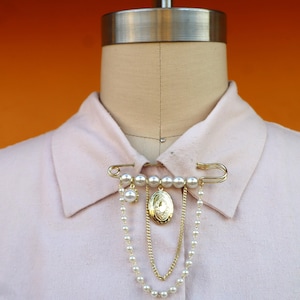 May include: A gold-toned safety pin brooch with faux pearls and a small locket. The brooch is attached to a light pink collared shirt on a mannequin. The locket hangs from a chain and is surrounded by faux pearls.