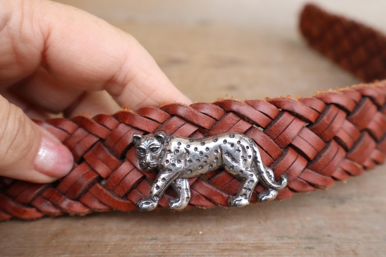 May include: A brown leather belt with a plaited design. The belt features a silver-coloured leopard-shaped buckle. The leopard has a spotted pattern and is in a walking pose. The belt is held by a person.