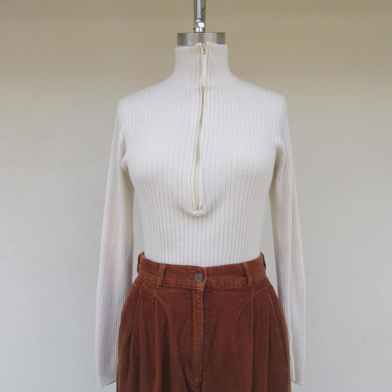 May include: A white ribbed turtleneck sweater with a zipper up the front. The sweater is on a mannequin and is paired with brown corduroy pants.