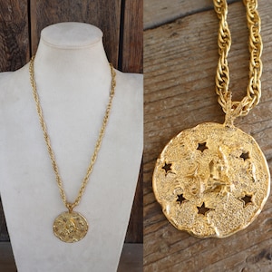 May include: A gold-toned necklace with a large, textured pendant. The pendant features a central figure and star cutouts. The chain is a twisted rope design, and the necklace is displayed on a mannequin.