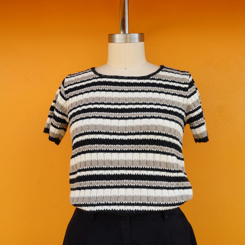 May include: A striped short-sleeved sweater in black, white and beige. The sweater is on a mannequin and has a round neckline.