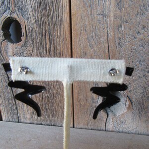 Pode incluir: Um par de brincos pretos de forma abstrata com postes prateados. Os brincos s&atilde;o exibidos em um suporte branco contra um fundo de madeira.
