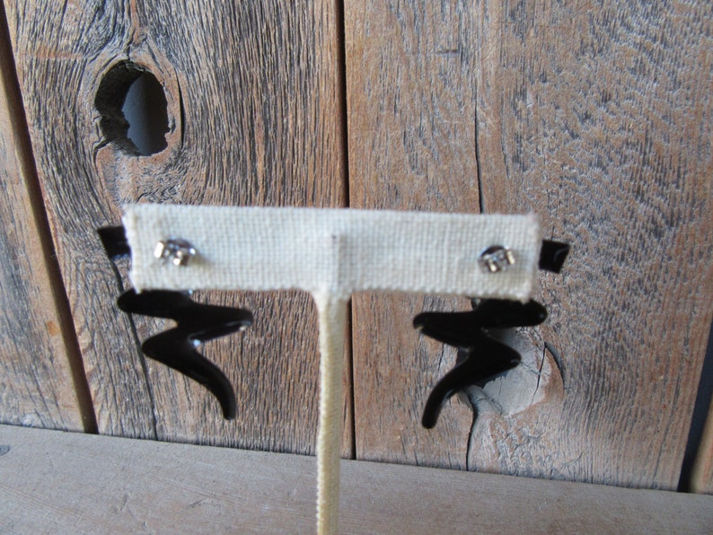 Pode incluir: Um par de brincos pretos de forma abstrata com postes prateados. Os brincos s&atilde;o exibidos em um suporte branco contra um fundo de madeira.
