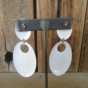 May include: A pair of white oval-shaped earrings with a smaller oval cutout in the center. The earrings are hanging from a metal stand.
