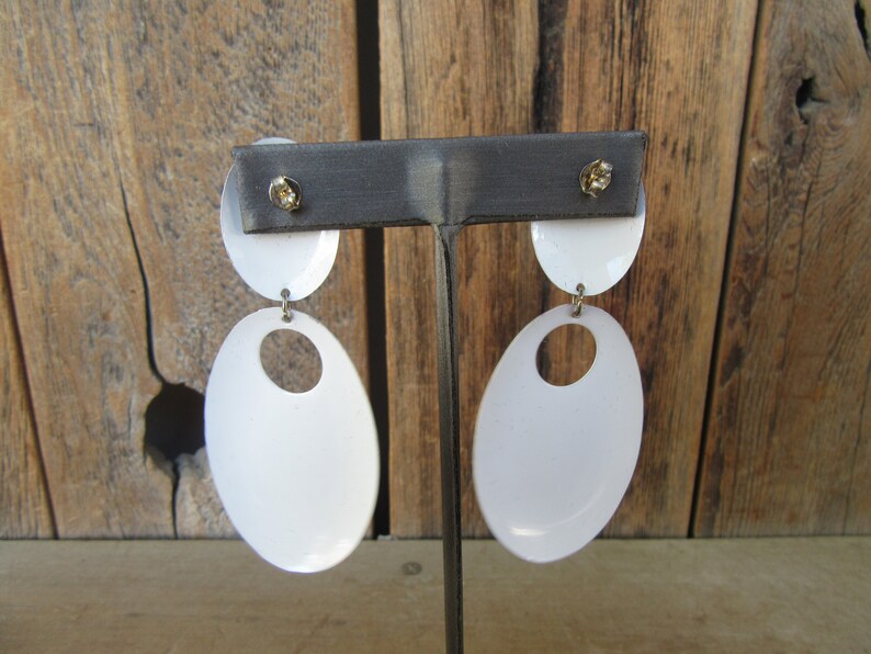 May include: A pair of white oval-shaped earrings with a smaller oval cutout in the center. The earrings are hanging from a metal stand.