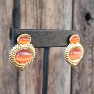 Può includere: Un paio di orecchini dorati con pietre ovali arancioni. Gli orecchini hanno un design testurizzato attorno alle pietre.