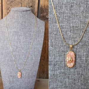 May include: A gold-toned necklace with a long chain and a pendant. The pendant is an oval shape with a pink stone and a gold diamond-shaped accent in the center. The necklace is displayed on a gray mannequin.