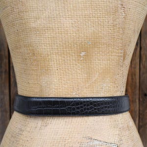May include: A black belt with a crocodile-like texture is wrapped around a mannequin. The belt is made of a dark material and has a rectangular buckle. The background is a wooden surface.